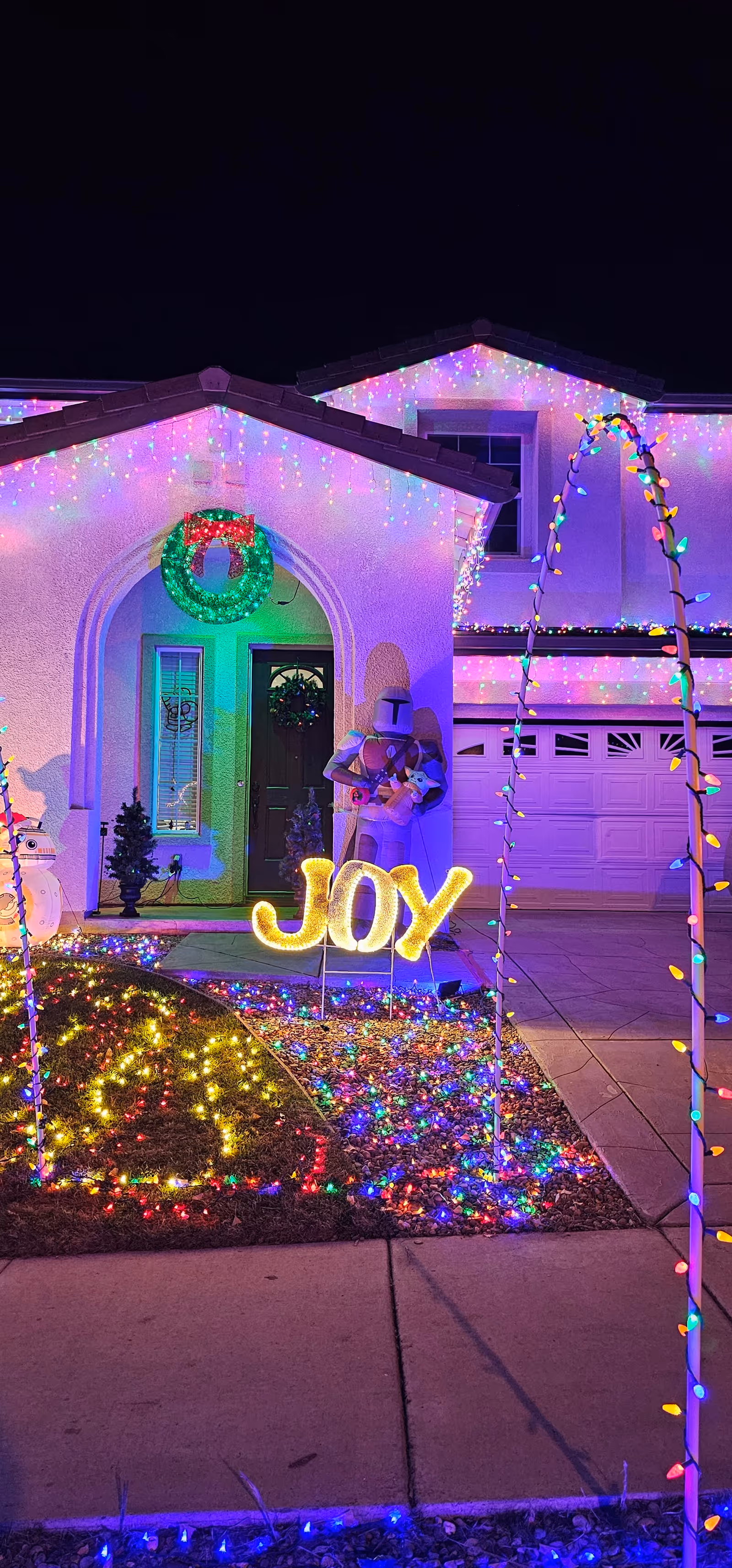 Front yard and entrance of a house lit with colorful holiday lights and JOY sign.