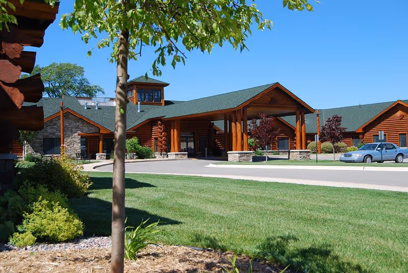 Exterior view of Freighter View Assisted Living facility showing a large log cabin style building with a green roof, a covered entrance supported by wooden pillars, a well-maintained lawn, small trees, shrubs, and a parked car on the driveway under a clear blue sky.