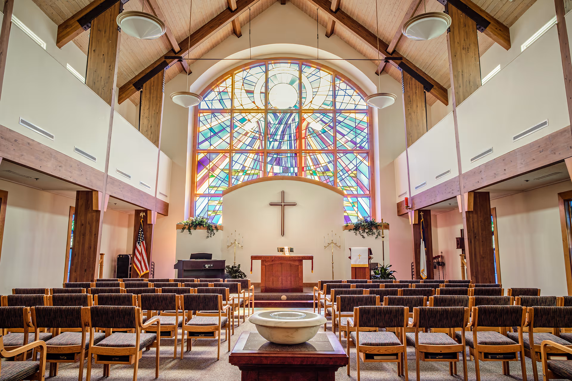 Interior of a chapel with rows of chairs facing an altar, a central cross, and a large stained-glass window.