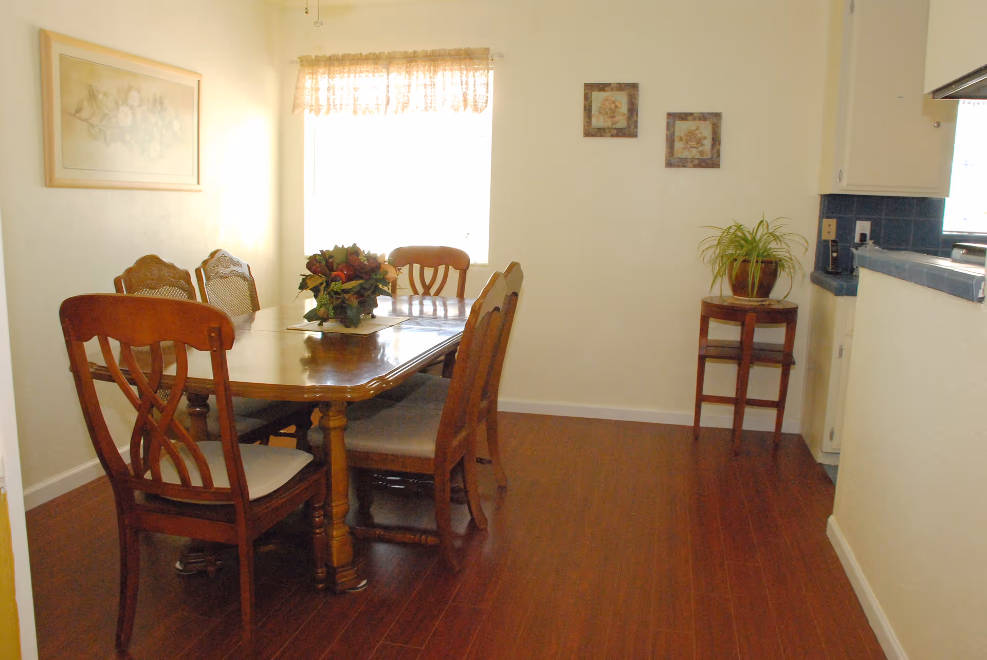 A dining room with a wooden dining table and six chairs. The table has a floral centerpiece. There is a window with a lace curtain, two framed pictures on the wall, and a small table with a potted plant near the kitchen counter.
