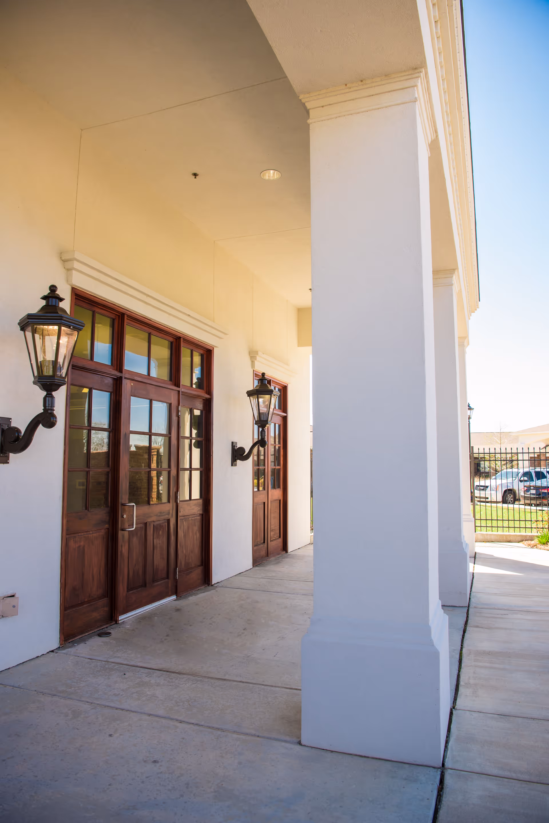 Exterior view of a building entrance with large white columns, wooden double doors with glass panes, and wall-mounted lantern-style lights on either side of the doors. A paved walkway runs alongside the building, and a fence and parked cars are visible in the background.