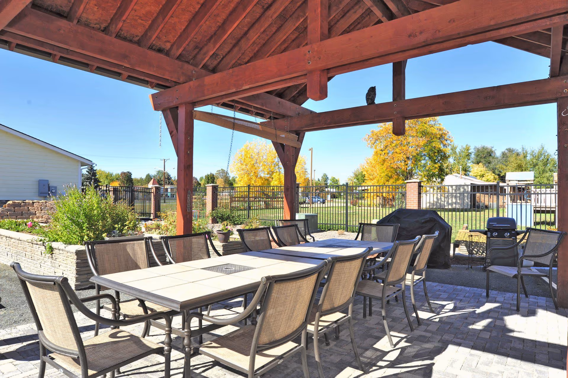 Outdoor covered patio area with a long rectangular table surrounded by multiple chairs. The patio has a wooden roof structure and is paved with bricks. There is a grill covered with a black cover and some greenery and trees visible in the background under a clear blue sky.