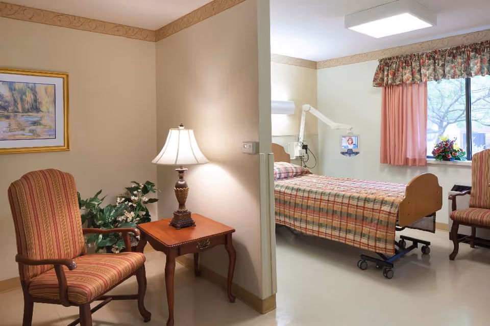 A cozy senior living facility room with a hospital-style bed covered in a plaid blanket, positioned near a window with floral curtains. The room has two striped armchairs, a wooden side table with a decorative lamp, a framed landscape painting on the wall, and a potted plant. A monitor arm is attached near the bed, displaying a small screen.
