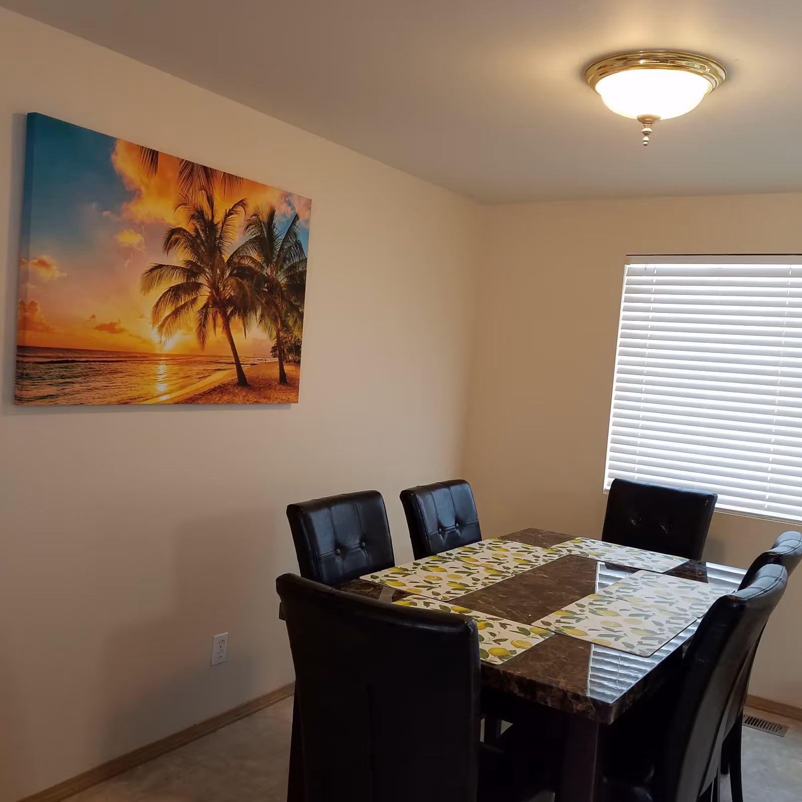 A dining room with a rectangular marble table surrounded by six black cushioned chairs. The table has four placemats with a lemon pattern. On the wall, there is a large colorful painting of a sunset beach scene with palm trees. A window with closed white blinds is visible, and a ceiling light fixture is turned on.