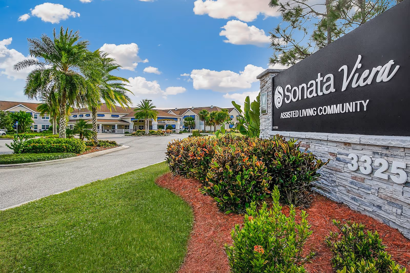 Entrance to Sonata Viera assisted living community with a large sign displaying the facility name and address, surrounded by well-maintained landscaping including bushes, palm trees, and green grass under a partly cloudy blue sky.