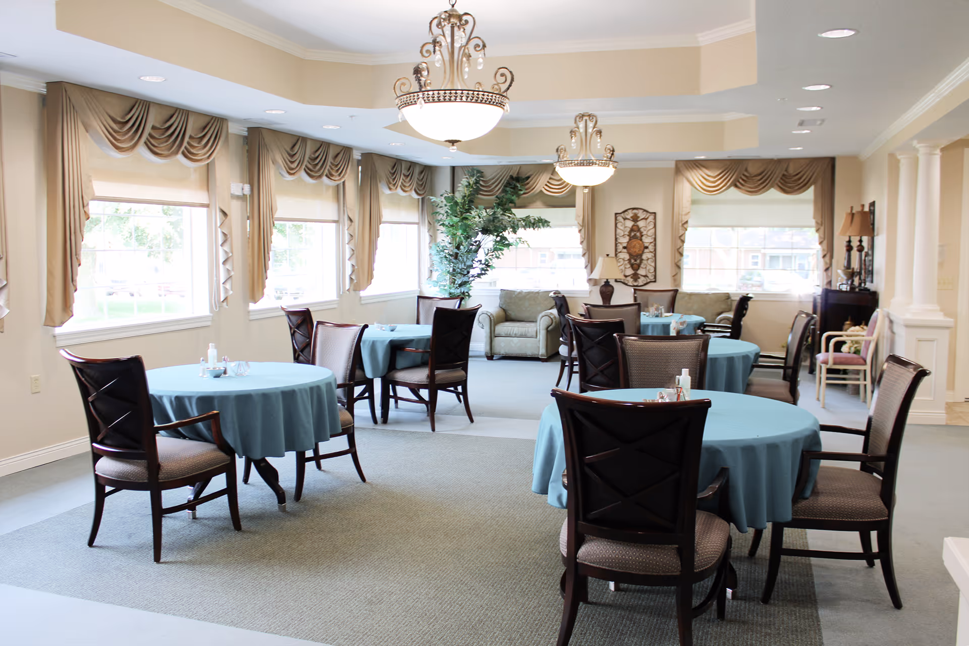 Bright dining room with round tables, blue tablecloths, and cushioned chairs.