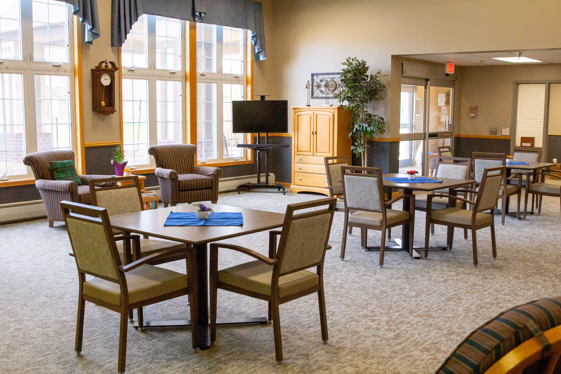 A spacious senior living lounge with multiple tables, chairs, and a TV in a common area.