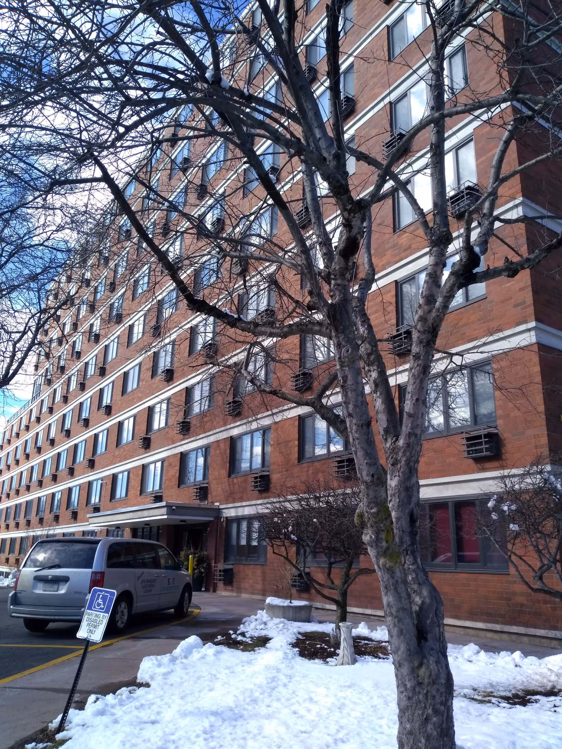 Multi-story brick senior living building exterior with parked van and bare trees in snow.