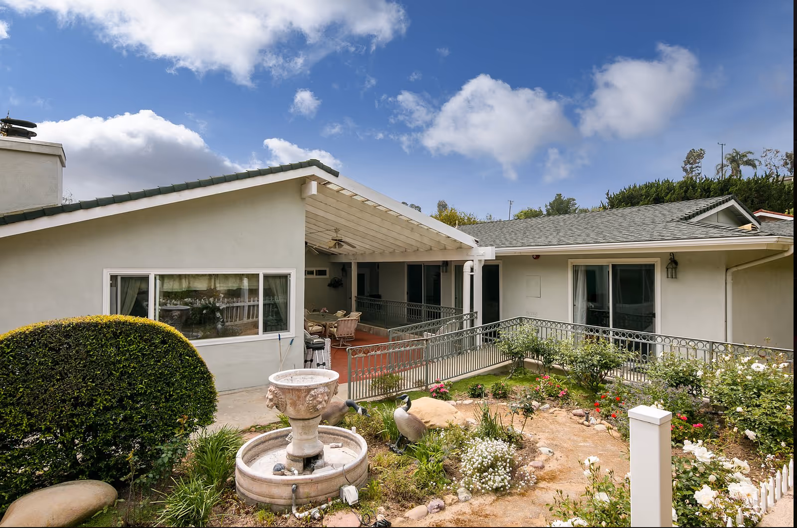 Front exterior of a single-story senior living cottage with a covered patio, metal railing and a landscaped garden with a fountain.