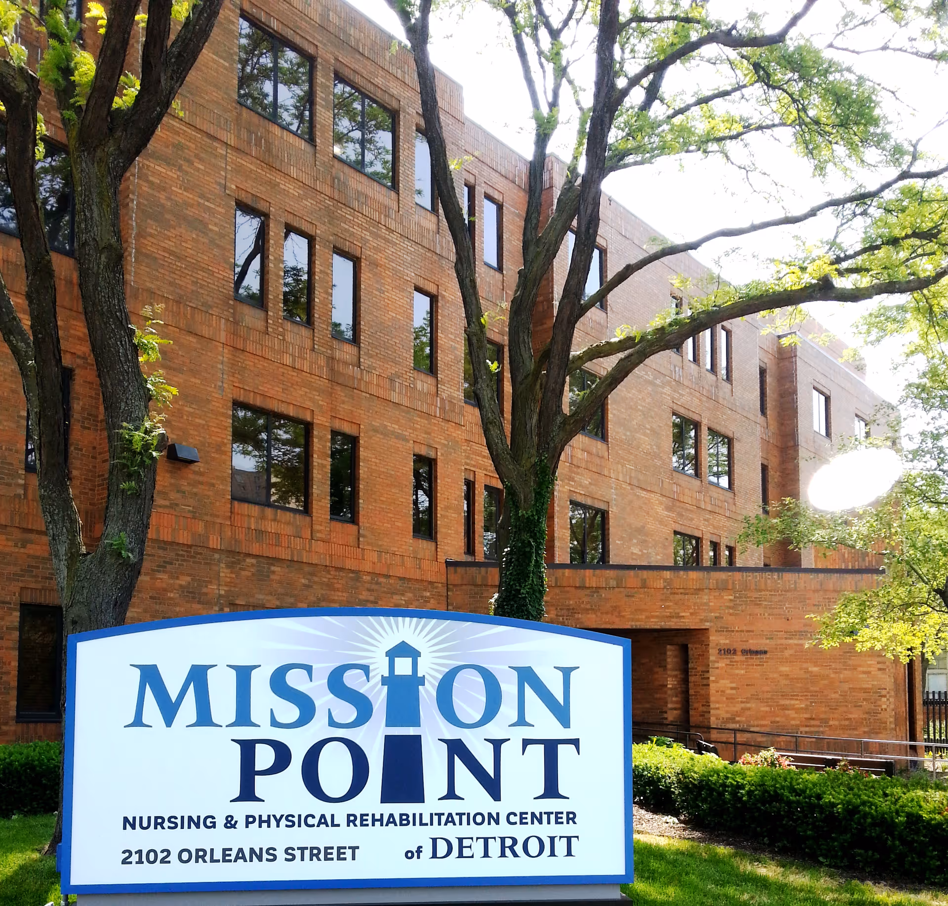 Exterior view of a multi-story brick building with several windows, partially shaded by large trees. In the foreground, there is a sign that reads 'Mission Point Nursing & Physical Rehabilitation Center, 2102 Orleans Street of Detroit.'