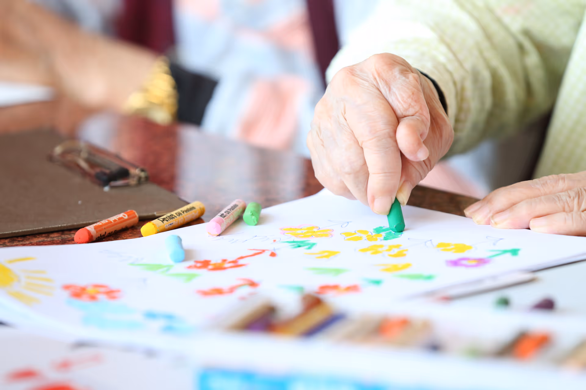 Close-up of elderly hands coloring with a green crayon on a colorful drawing.
