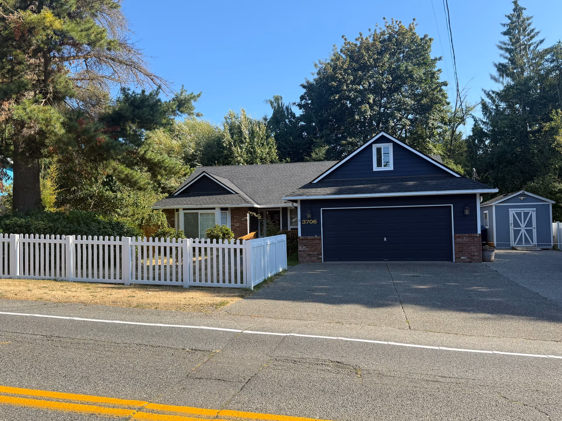 Blue single-story house with a two-car garage, white fence, and trees in the front yard.