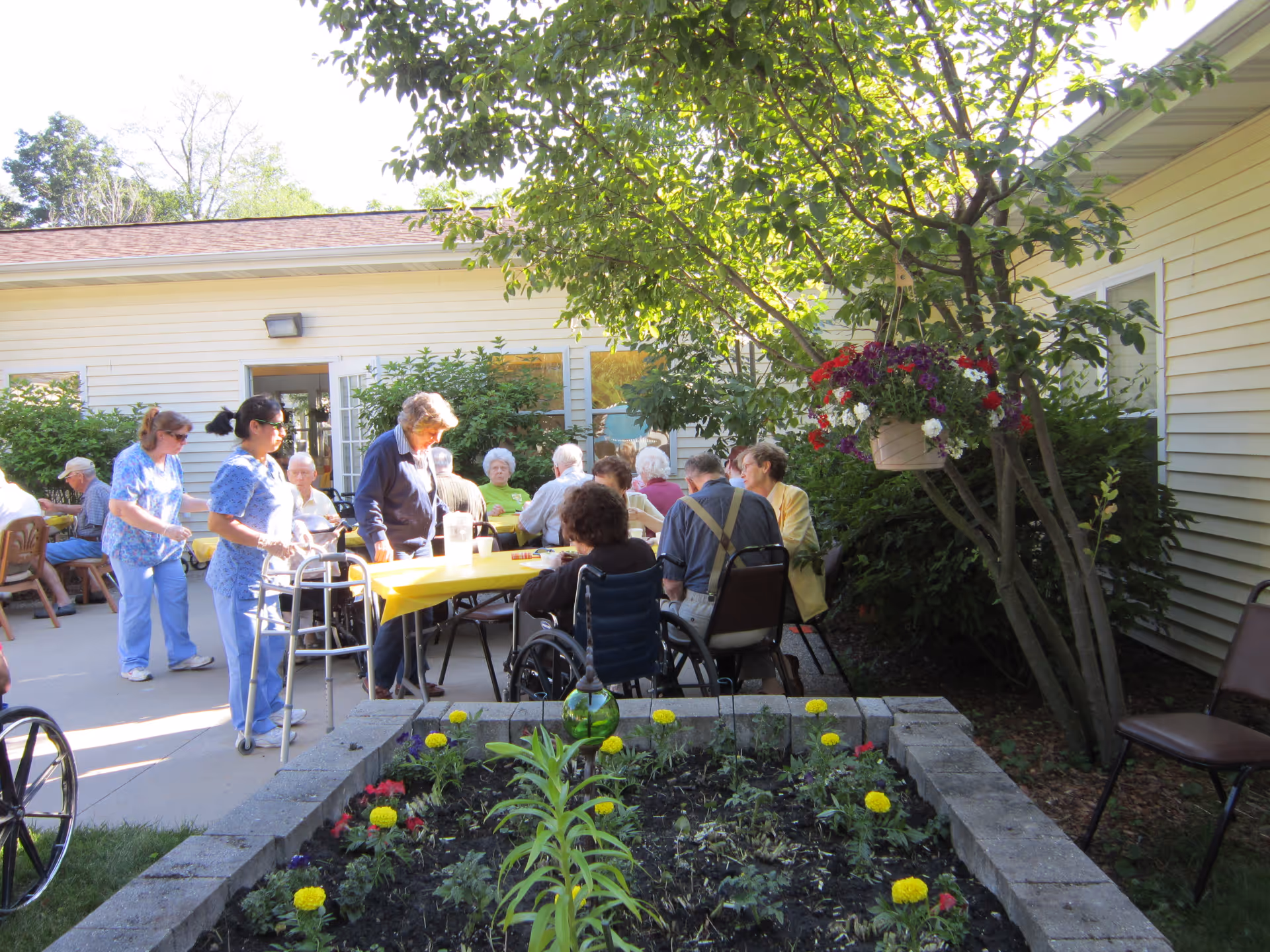 Seniors and caregivers gathered around a table in an outdoor courtyard garden at an assisted living facility.