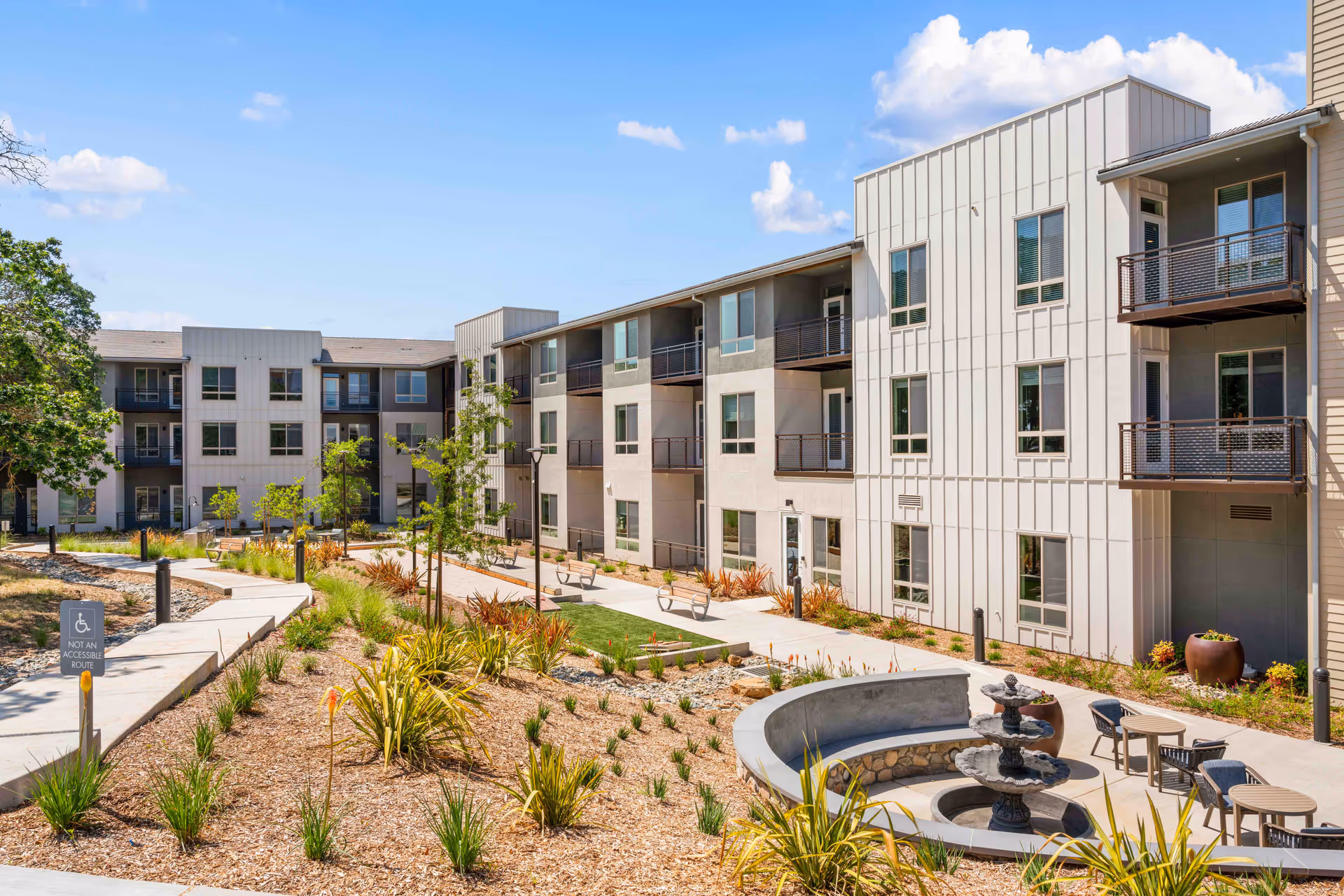 Three-story modern senior living building surrounding a landscaped courtyard with walkways, benches, and a fountain.