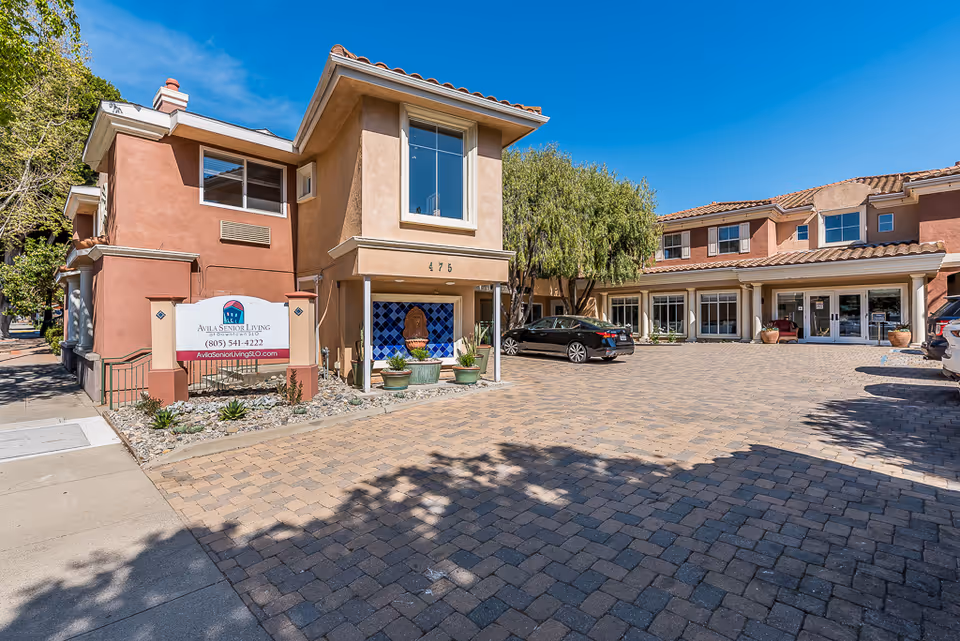 Exterior view of Avila Senior Living at Downtown SLO showing a two-story building with a paved driveway and parked cars under a clear blue sky. There is a sign with the facility's name, phone number, and website near the sidewalk.