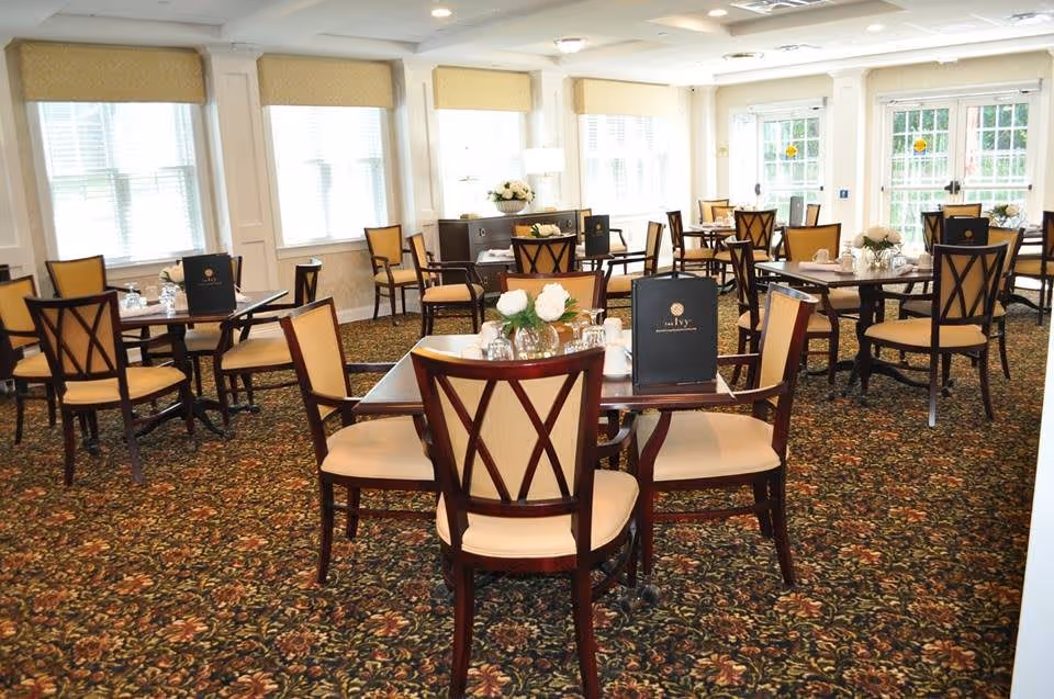 A bright dining room with multiple wooden tables and chairs with beige cushions. Each table is set with menus, glassware, and small floral centerpieces. Large windows with beige valances let in natural light, and a patterned carpet covers the floor.