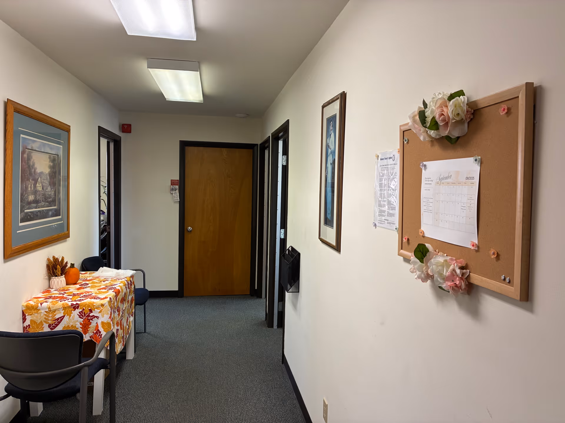 Interior hallway of a senior living facility with a corkboard, flowers, and a table along wall.