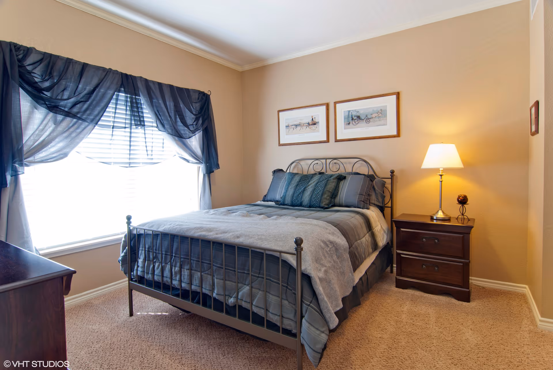 Neat bedroom with a metal-framed bed, bedside table and lamp, large window with dark drapes, and framed art on beige walls.