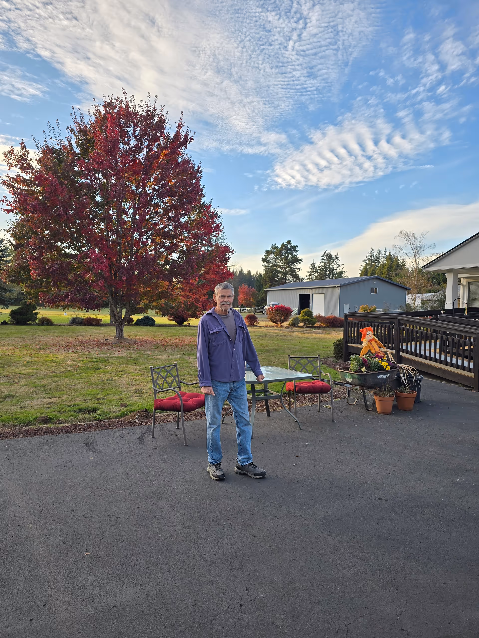 Older man stands on a paved outdoor patio with furniture, autumn tree, and building in background.