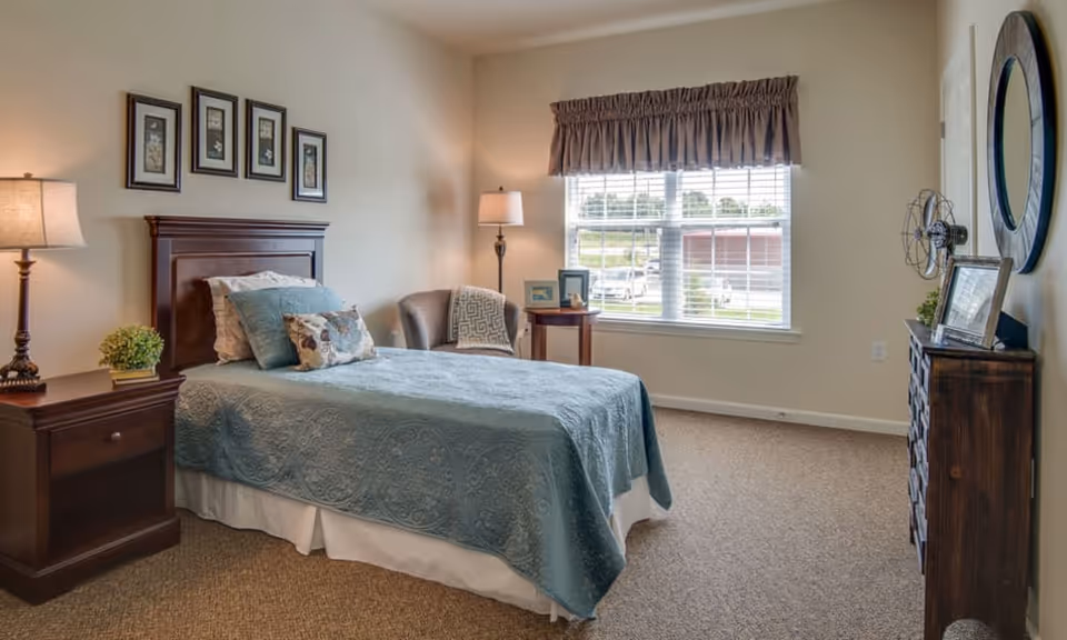 A cozy bedroom in a senior living facility featuring a single bed with a blue quilt and decorative pillows, a wooden nightstand with a lamp and a small plant, a comfortable armchair with a throw blanket, a round side table with framed photos, a window with blinds and a valance, and a wooden dresser with a round mirror and decorative items on top.