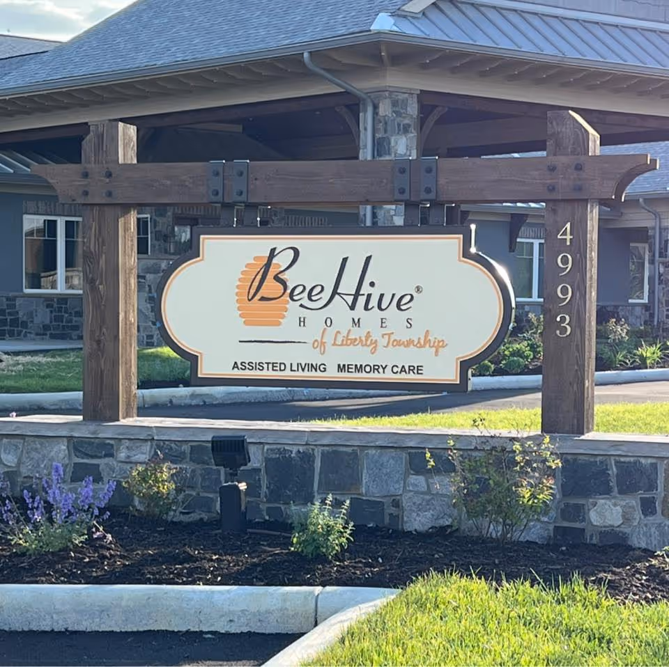 Outdoor view of a wooden sign for Beehive Homes of Liberty Township, an assisted living and memory care facility, mounted on a stone base with a building and landscaping in the background.