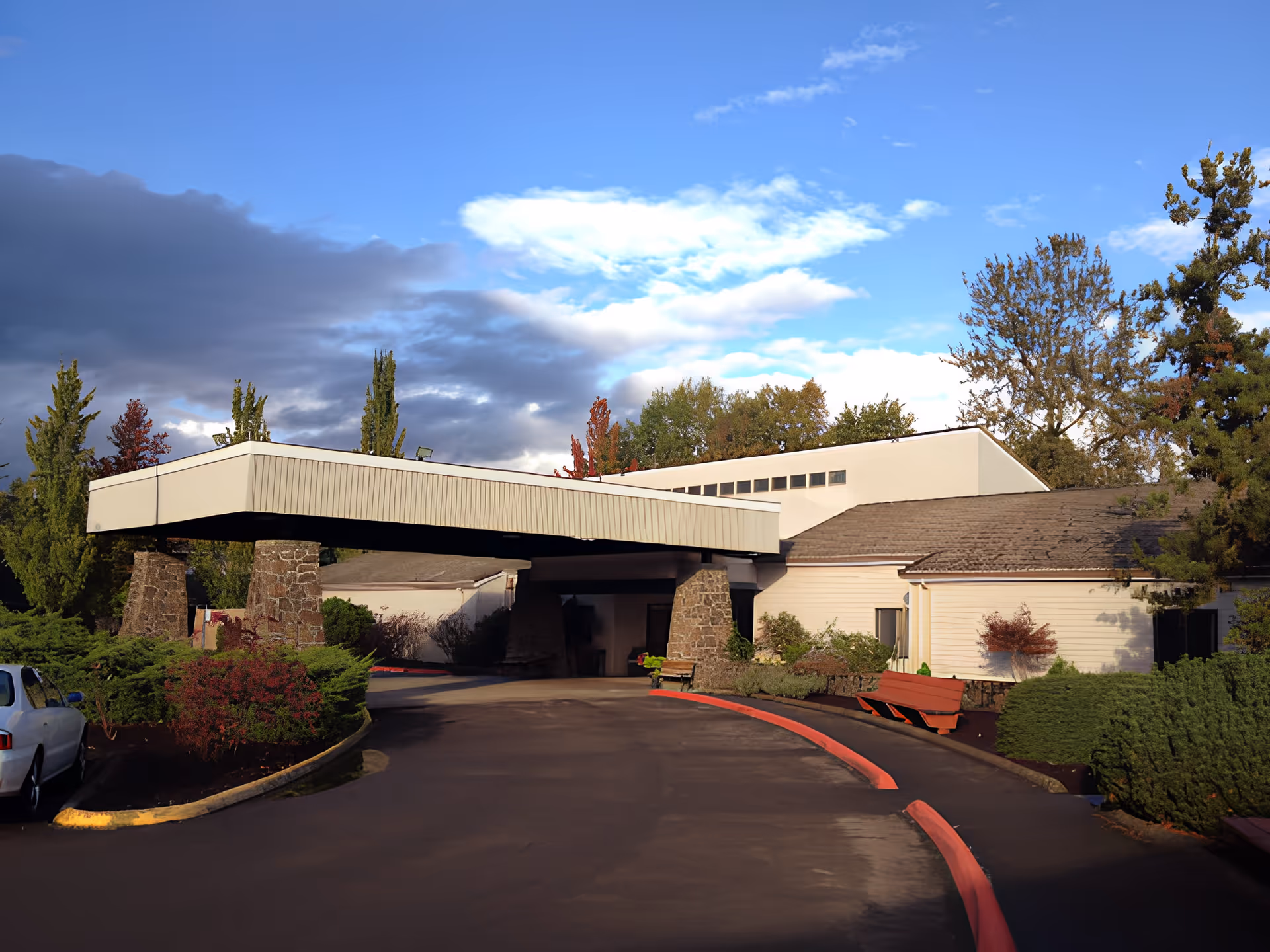 Exterior view of Churchill Estates Retirement Community building entrance with a covered driveway supported by stone pillars, surrounded by trees and bushes under a partly cloudy sky.