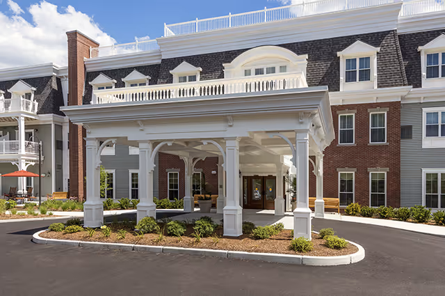 Entrance of a senior living facility with a large white covered porte-cochere supported by columns, surrounded by landscaped greenery and a paved driveway. The building behind has multiple windows and a combination of brick and siding exterior under a partly cloudy sky.