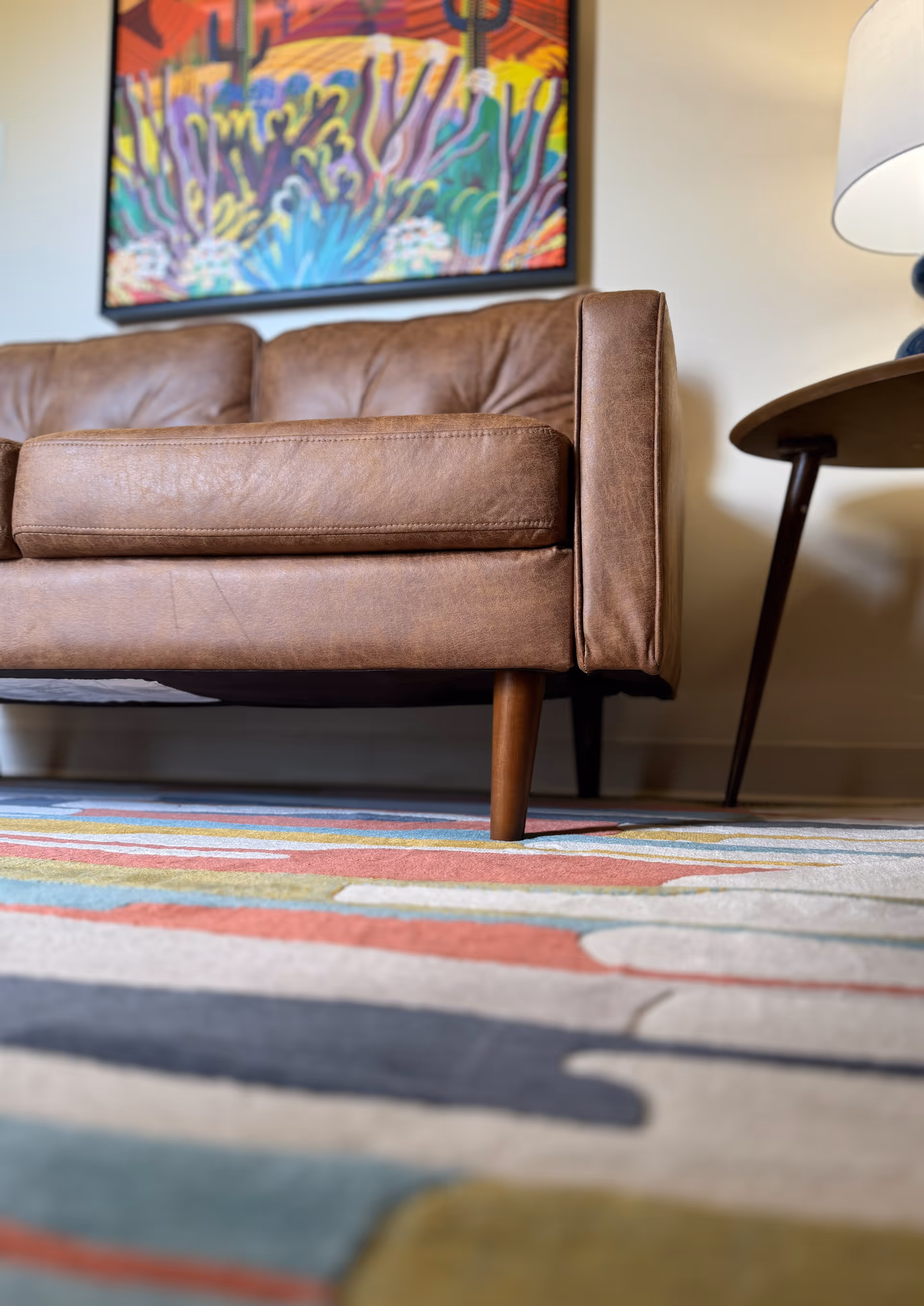 Close-up view of a brown leather sofa with wooden legs on a colorful patterned rug, a round wooden side table with a lamp, and a vibrant desert-themed painting on the wall in the background.