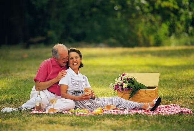 An elderly couple enjoying a picnic on a red and white checkered blanket in a grassy outdoor area. The man in a pink shirt is sitting behind the woman in a white shirt and checkered overalls, holding her affectionately. There is a picnic basket with flowers and food items around them.