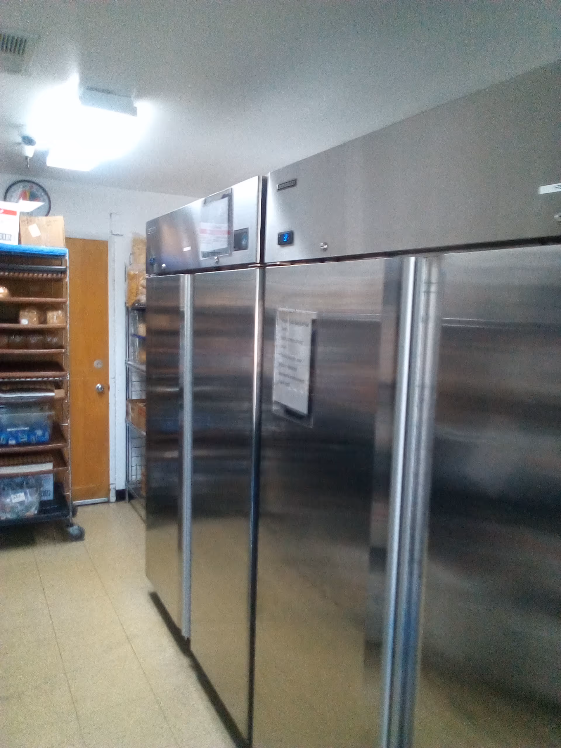 Industrial stainless steel refrigerators line a small kitchen area with shelves and a wooden door.