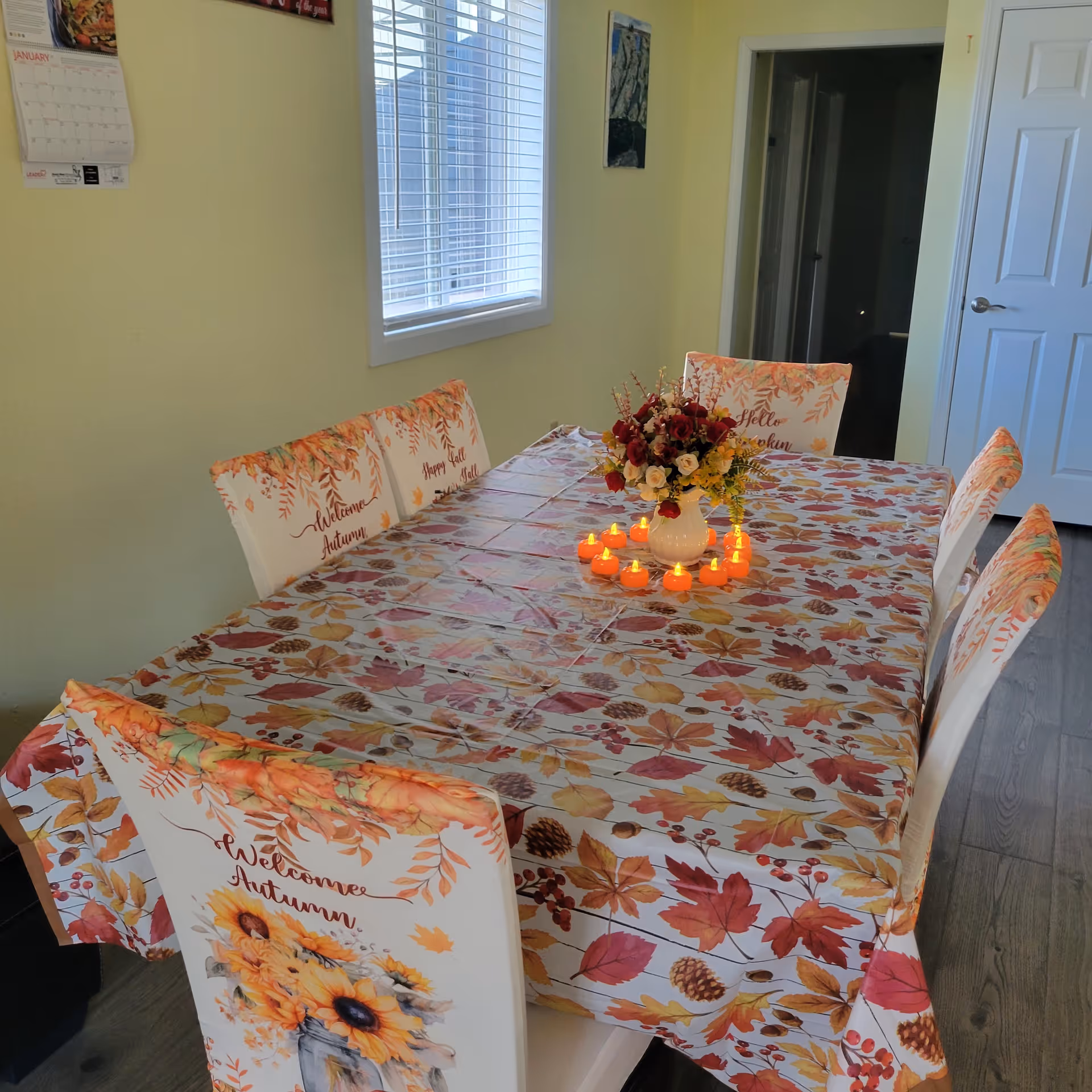 A dining table set for autumn with a tablecloth and chair covers featuring fall leaves and sunflowers. The table has a centerpiece of a vase with red and yellow flowers surrounded by small orange LED candles. The room has light yellow walls, a window with blinds, a calendar on the wall, and a white door in the background.