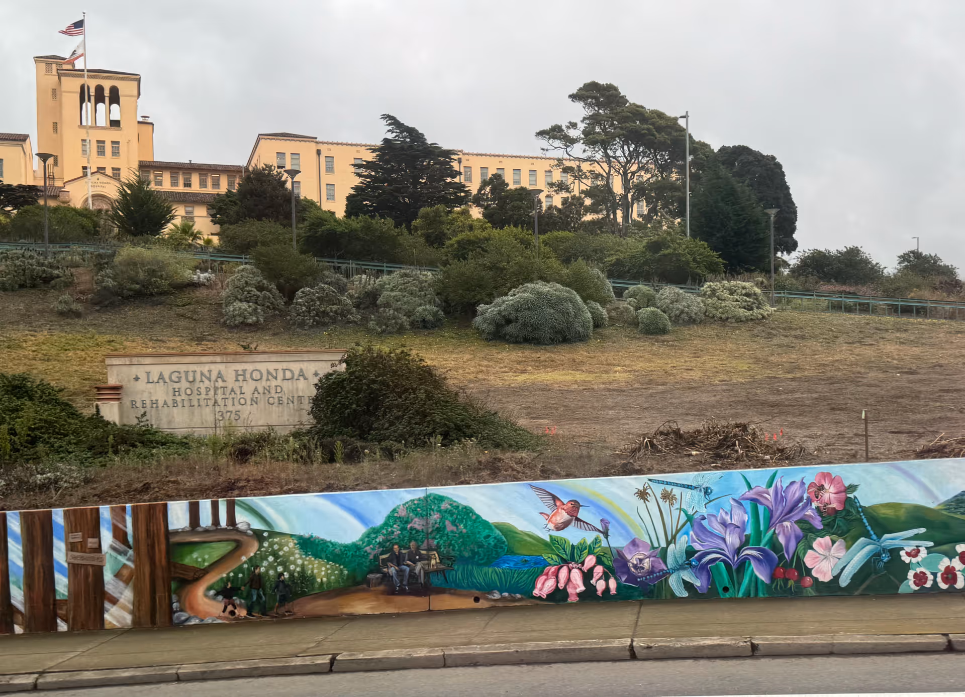 Laguna Honda Hospital exterior on a hillside with a colorful mural fence along street.