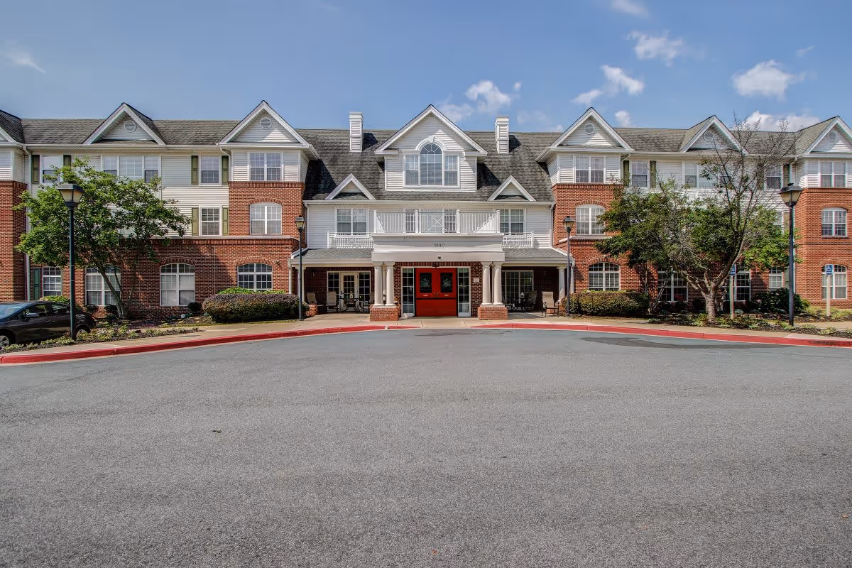 Front exterior view of a senior living facility building with a red double-door entrance, white columns, and a combination of brick and white siding. The building has multiple windows, a pitched roof, and is surrounded by trees and shrubs under a partly cloudy sky.