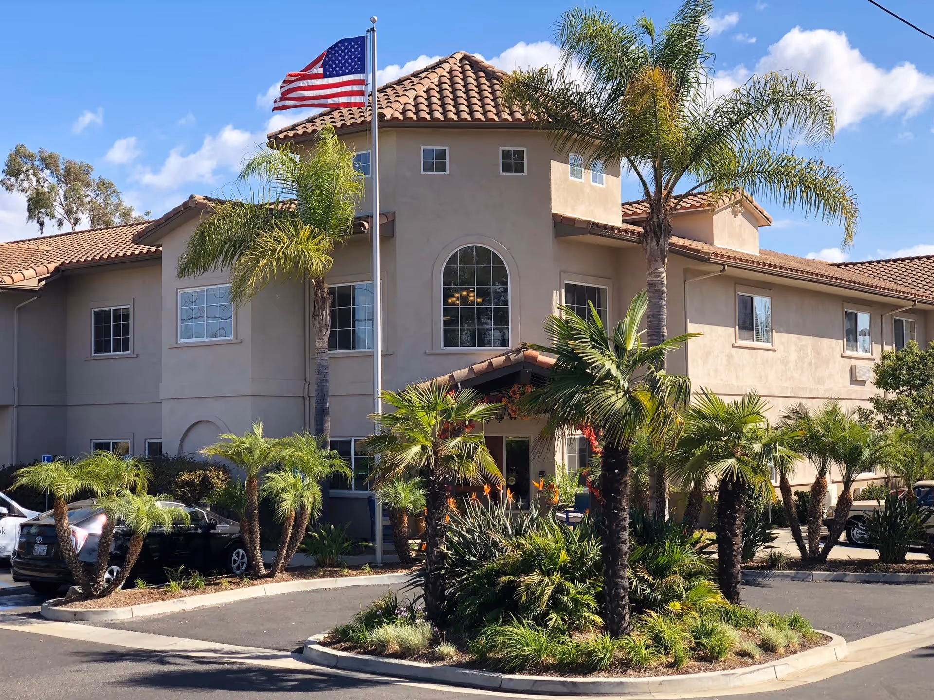Two-story beige Regency Fallbrook building with palm trees, flag, and manicured landscaping.