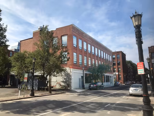 Brick multi-story building on a city street with large windows and surrounding trees.