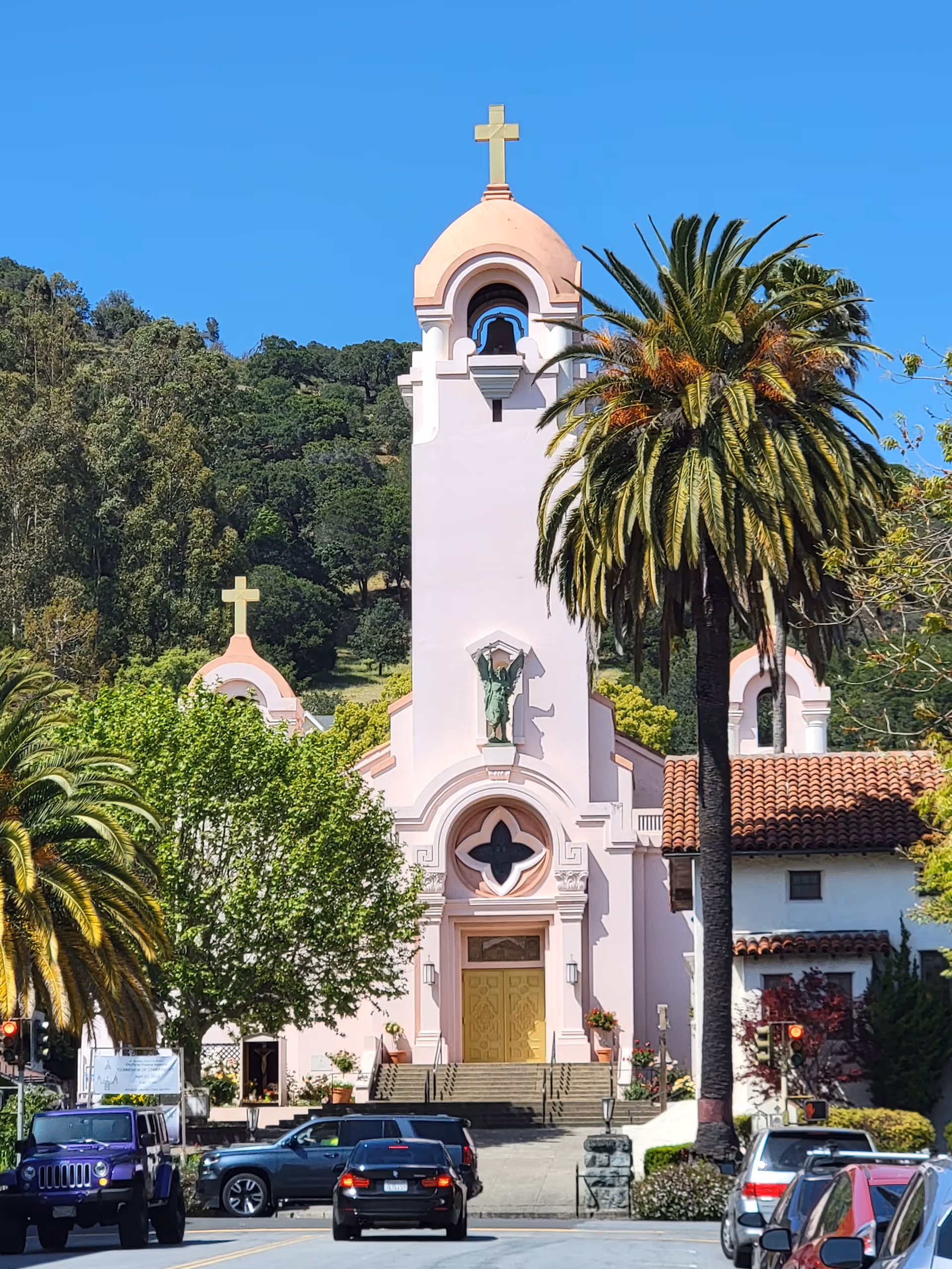 Facade of a pink church with a bell tower, cross, palm trees, and parked cars.