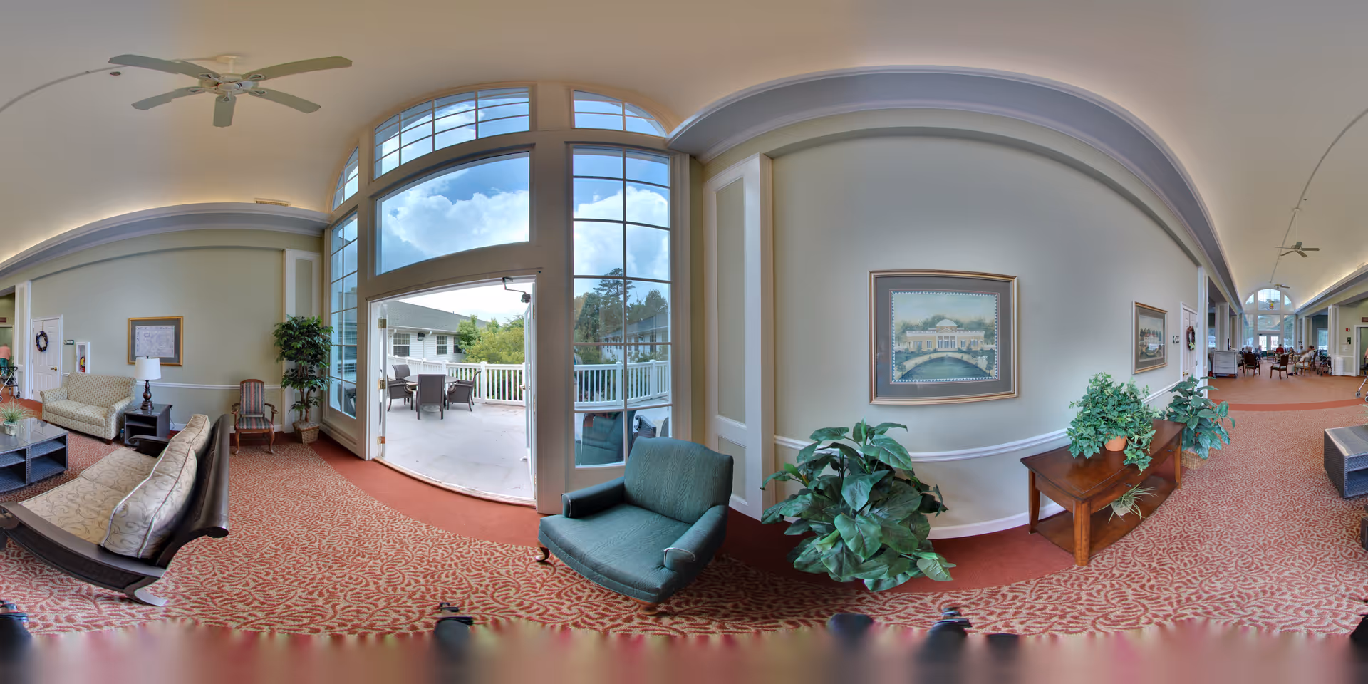 A spacious senior living facility common area with patterned red carpet, beige walls, and large windows letting in natural light. The room features comfortable seating including sofas and armchairs, a ceiling fan, potted plants, framed artwork on the walls, and a door leading to an outdoor patio with tables and chairs. The hallway extends into a dining area in the background.