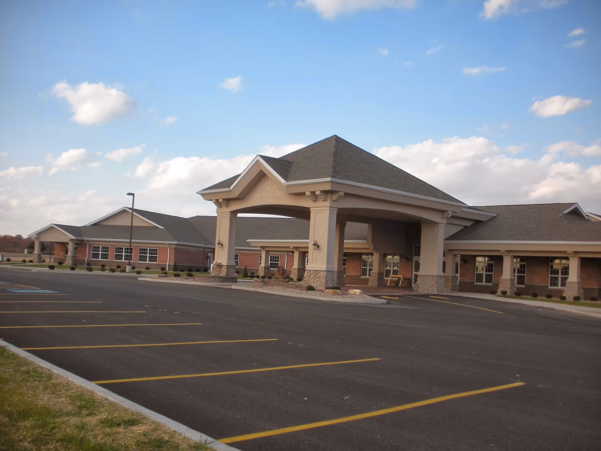 Exterior view of a single-story senior living facility building with a covered entrance and a large empty parking lot in front under a partly cloudy sky.