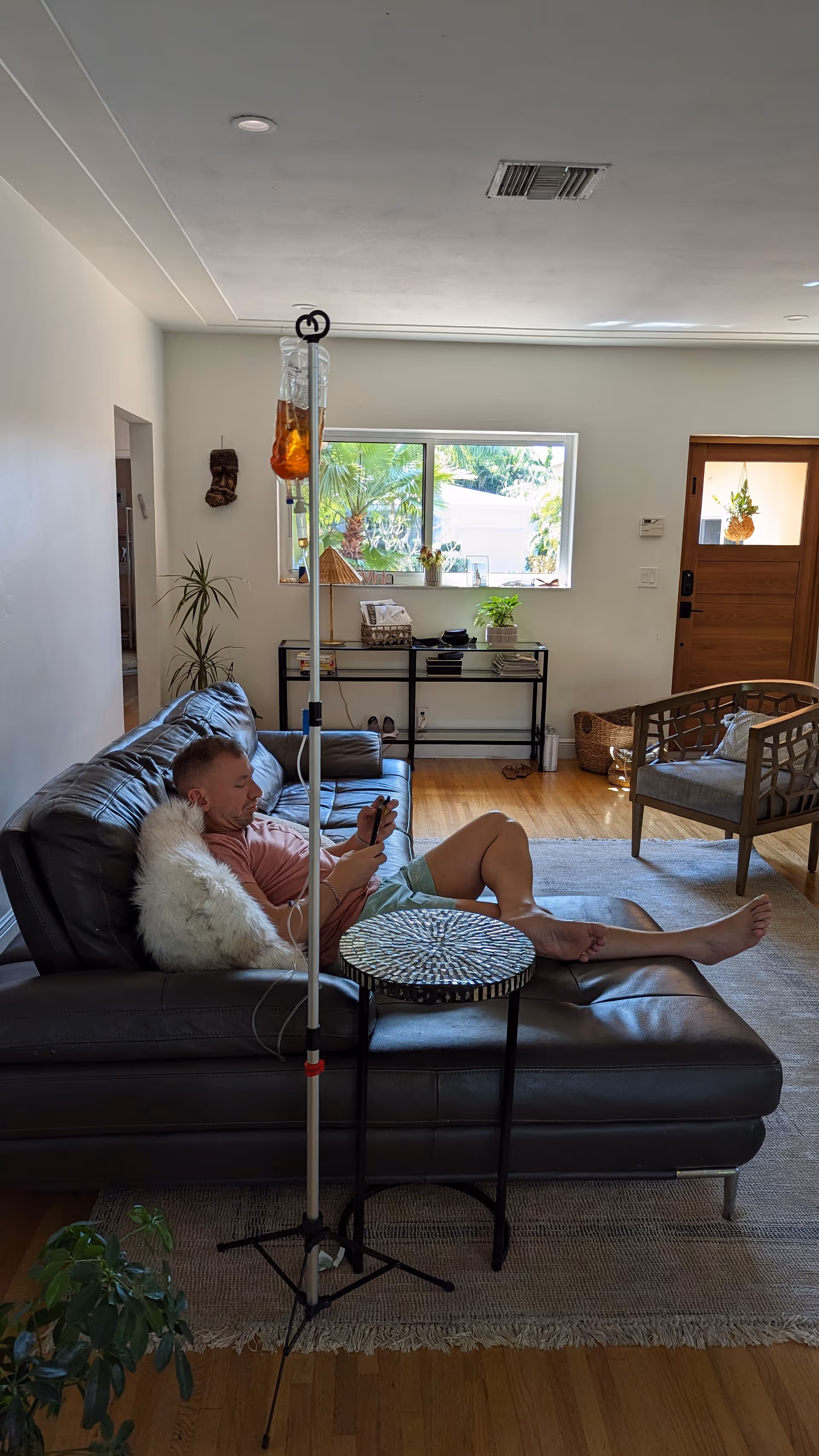 Resident lounges on a dark leather sofa with an IV drip in a bright living room.