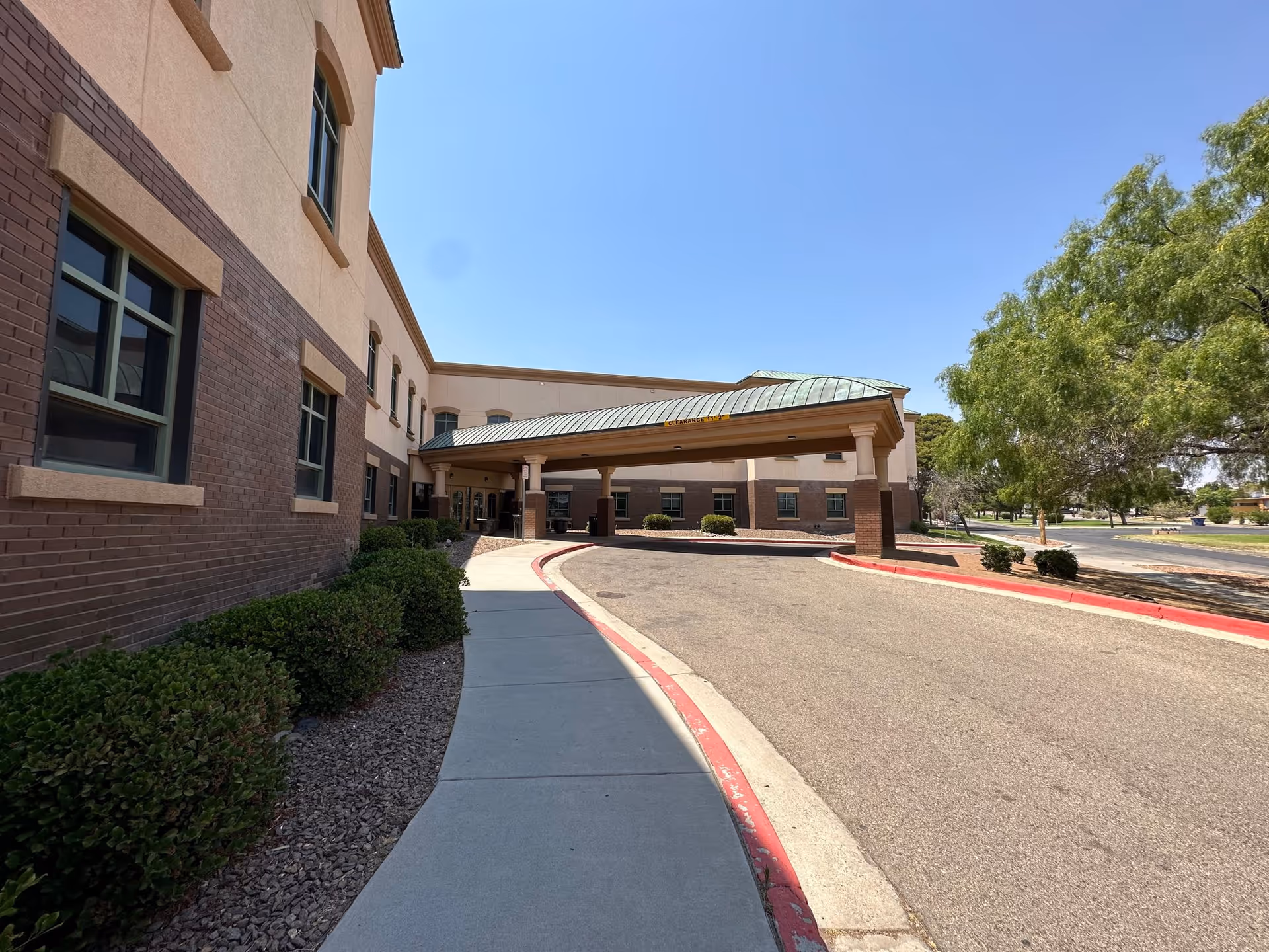 Exterior view of Nazareth Living Care Center showing a two-story building with a covered entrance driveway, bushes along the sidewalk, and trees on the right side under a clear blue sky.