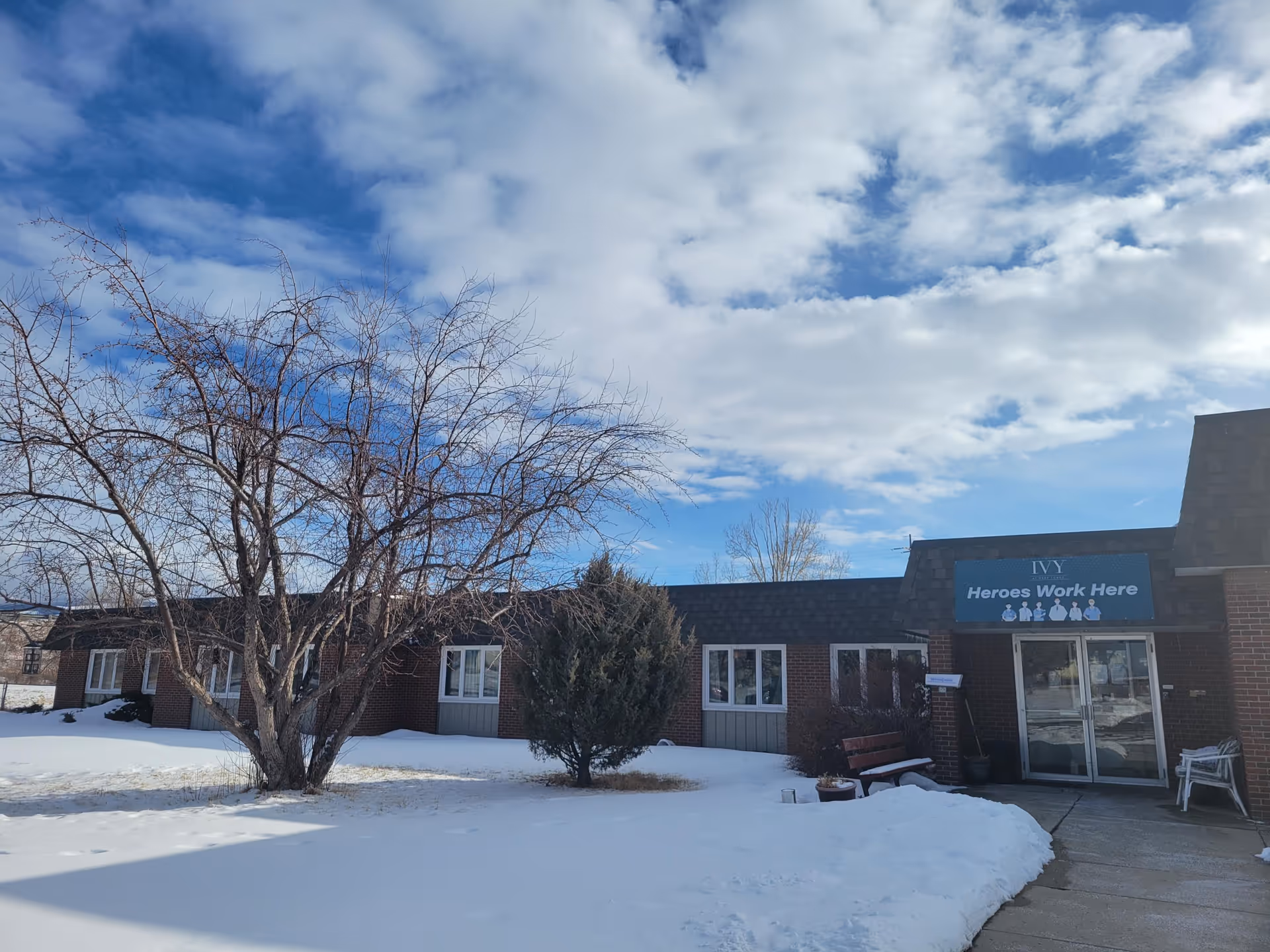 Brick senior living facility exterior with signage, snow-covered ground, leafless trees, and blue sky.