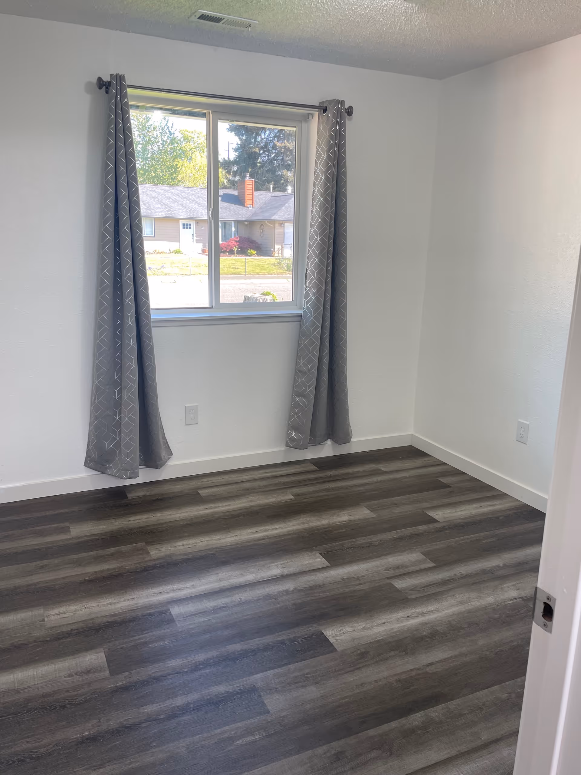Empty interior room with a window, gray curtains, and dark laminate flooring.