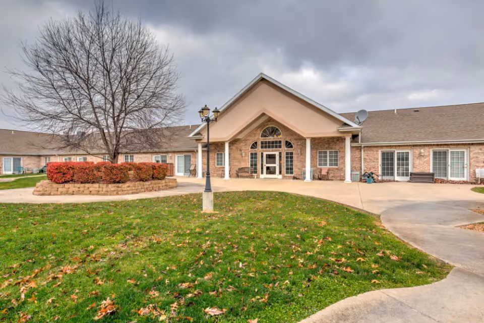 Front entrance of a single-story brick senior living building with a covered portico, lamppost, landscaped shrubs and a lawn under a cloudy sky.