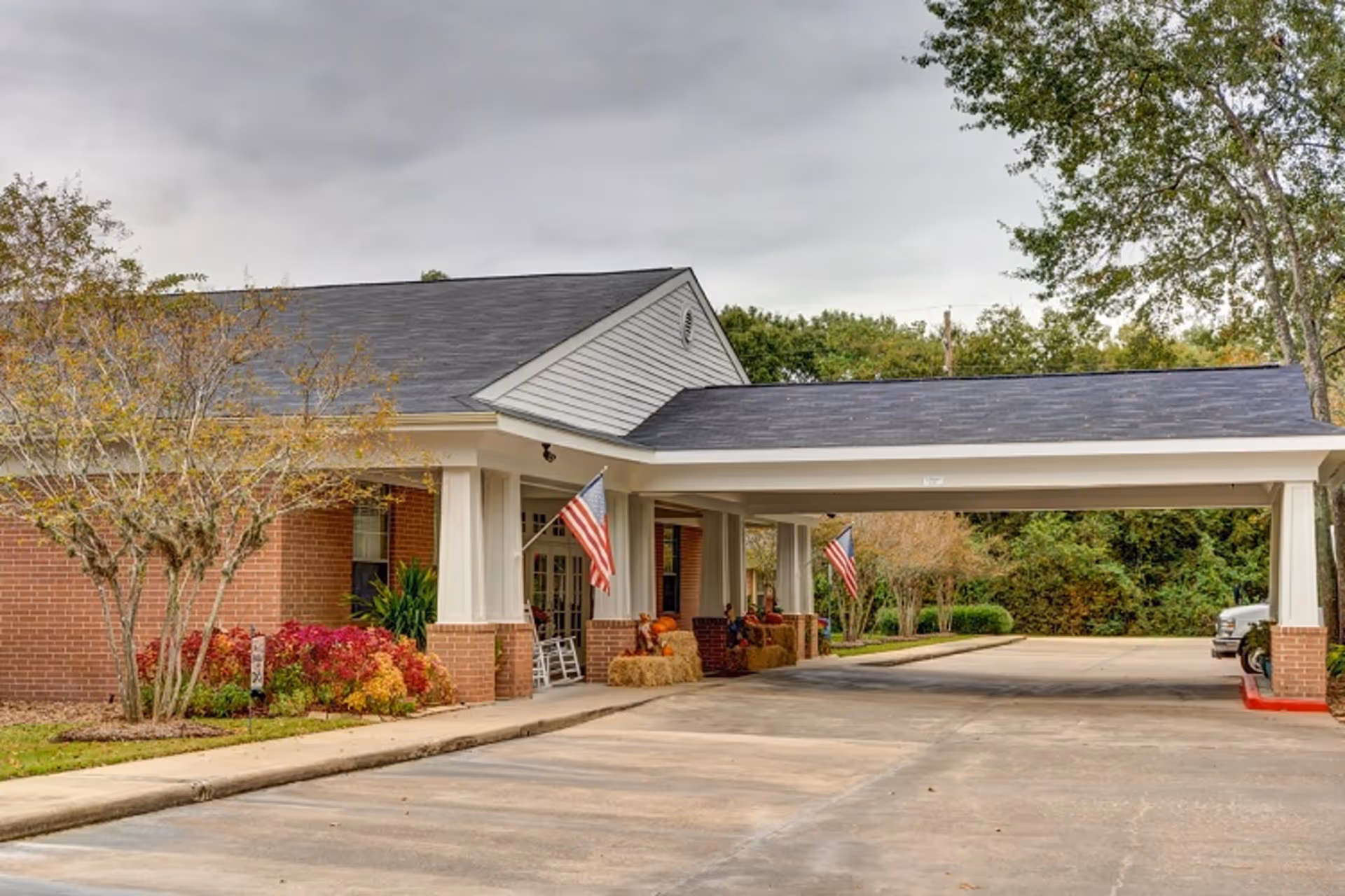 Exterior view of a senior living facility entrance with a covered drop-off area supported by white columns. The building has a brick facade with white siding above and two American flags displayed near the entrance. There are trees and shrubs around the building and a paved driveway leading up to the entrance.