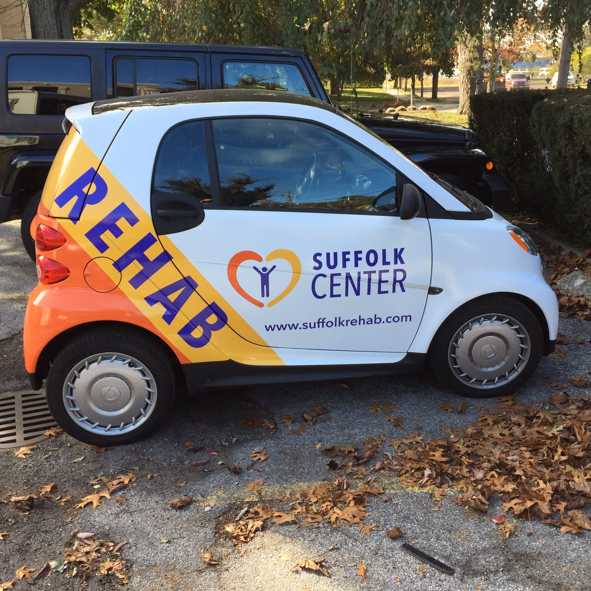 A small white and orange Smart car parked outdoors with the words 'REHAB' in large blue letters on the rear side and 'SUFFOLK CENTER' along with a heart-shaped logo and the website www.suffolkrehab.com on the door.