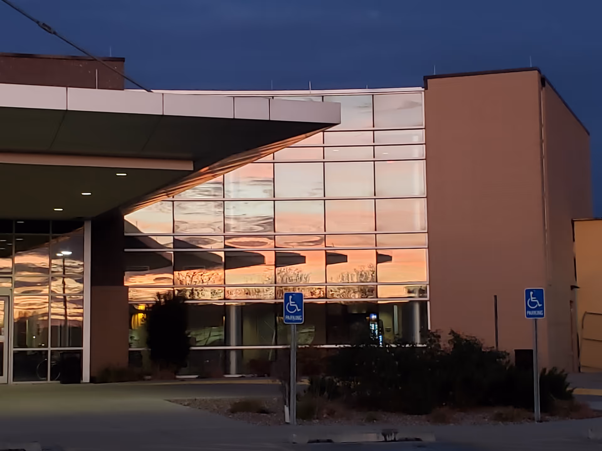 Modern building exterior with reflective glass facade showing sunset colors and handicap parking signs.