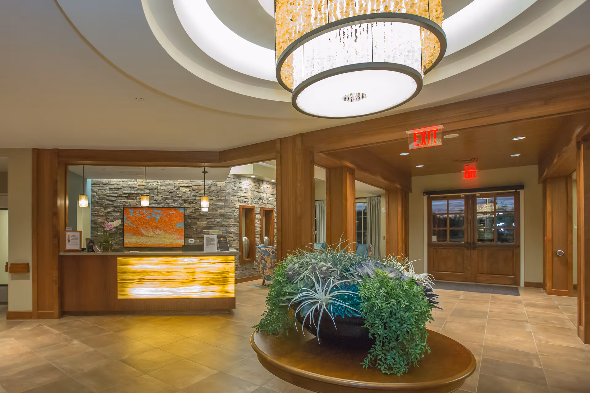 Interior view of a senior living facility lobby with a round wooden table holding a large plant arrangement in the center. Behind the table is a reception desk with a glowing front panel, stone accent wall, and hanging pendant lights. Wooden double doors with glass panels and exit signs are visible in the background. The ceiling features a large circular light fixture.