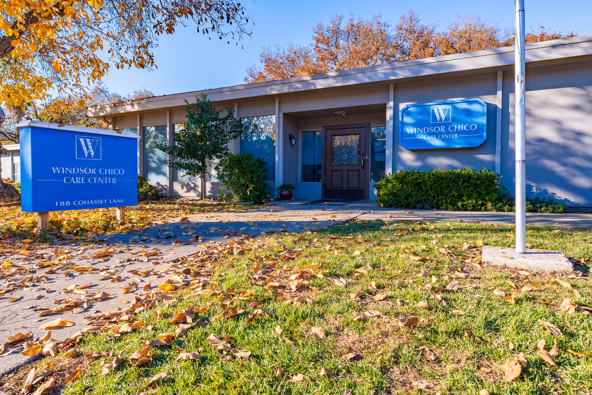 Exterior view of Windsor Chico Care Center building with a blue sign displaying the facility name and address. The building is surrounded by trees with autumn leaves on the ground and a clear blue sky.