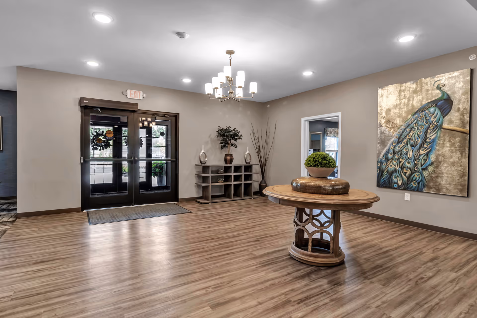 Interior view of a senior living facility entrance area with double glass doors, a round wooden table with a decorative plant centerpiece, a wooden shelf with decorative items, a large painting of a peacock on the wall, and a chandelier hanging from the ceiling.