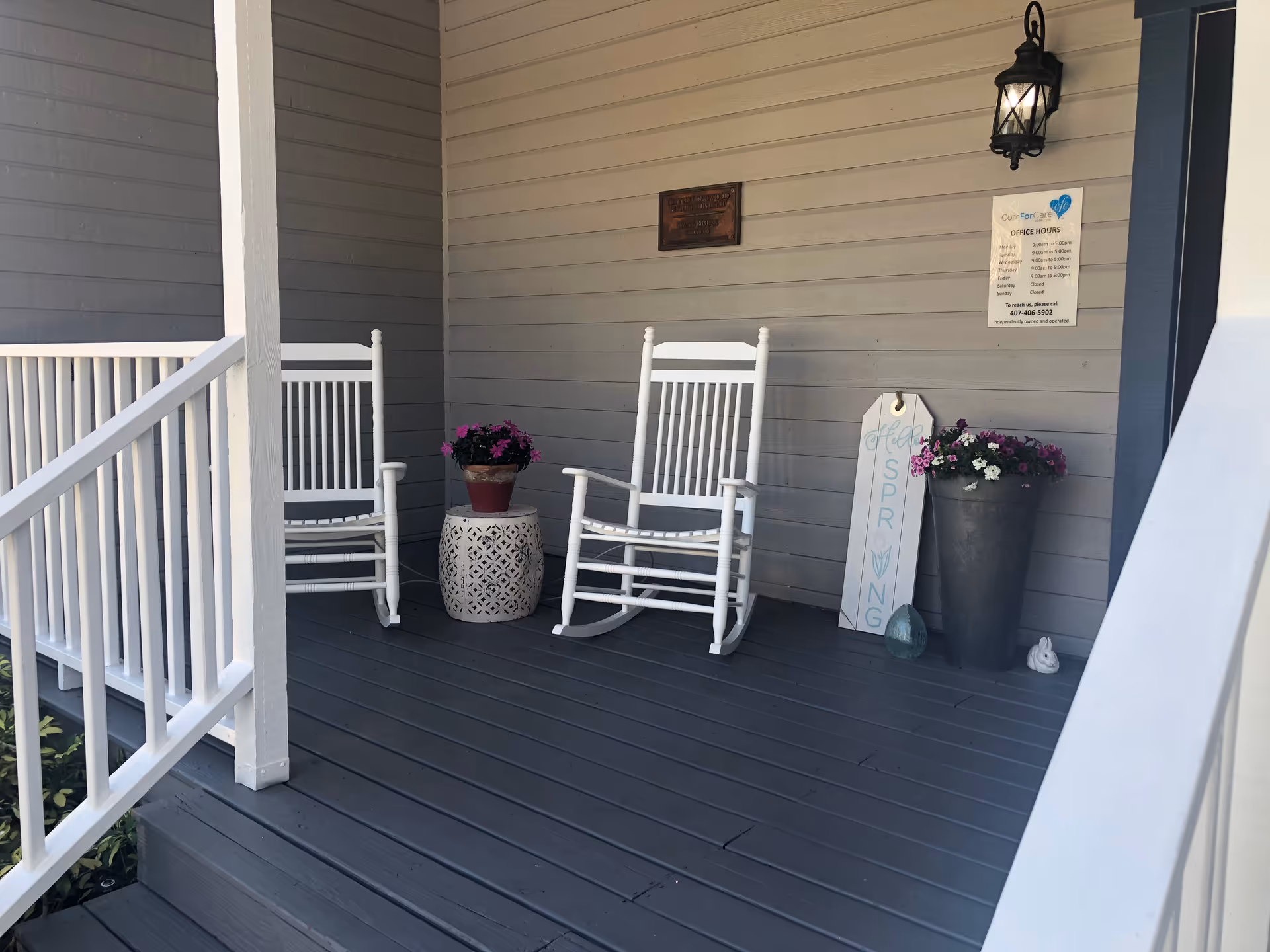 Porch with white rocking chairs, potted flowers, and decorative sign on gray siding.